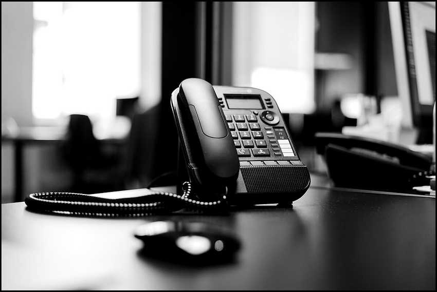 A black office desk phone on a desk, blurred office in the background, monochrome with high contrast